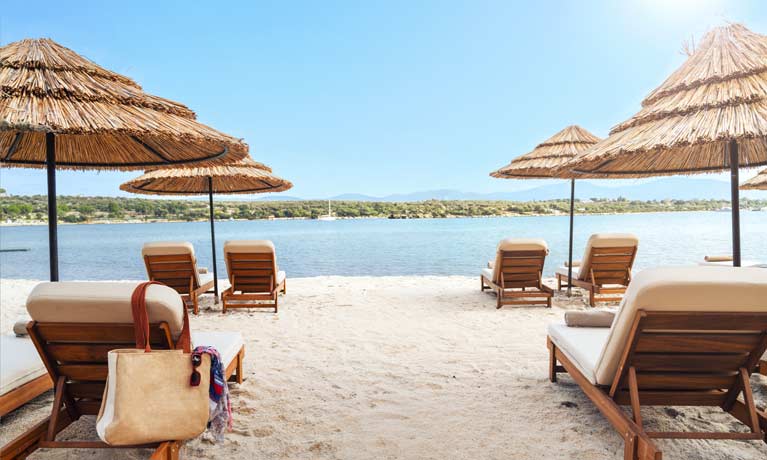 An inviting calm blue sea can be seen from between two sun loungers on the soft white sand, under the shade of a large umbrella