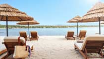 An inviting calm blue sea can be seen from between two sun loungers on the soft white sand, under the shade of a large umbrella