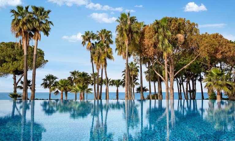 A large still blue outdoor swimming pool with a line of trees and the beach and sea behind