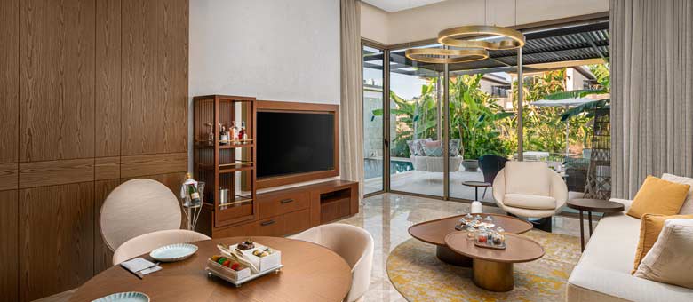 The lounge area inside one of the Bayou Villas, a large dining table and comfy sofa facing a large wall mounted television can be seen with a spacious patio and swimming pool beyond the floor to ceiling sliding glass doors
