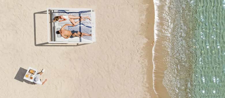 A couple stretch out on a day bed at the beach as a waiter approaches with a tray of refreshments.