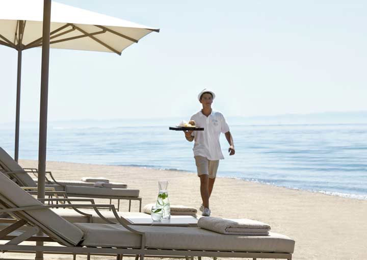 A smart yet casually dressed member of staff attends to guests needs while they relax on the beach, an example of the impeccable standards of service that is known as the 'ikos Concept'.