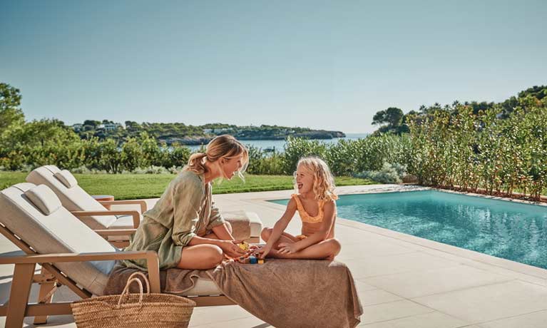 A mother and daughter enjoy quality time next to a still blue swimming pool with lovely views over countryside behind them.