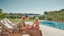 A mother and daughter enjoy quality time next to a still blue swimming pool with lovely views over countryside behind them.