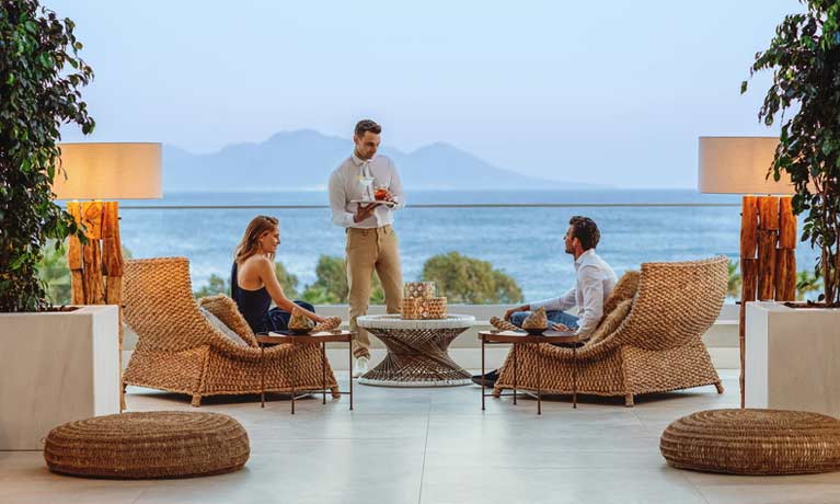 A couple relax at the Lobby Bar in front of beautful sea views while being served by a smiling waiter.