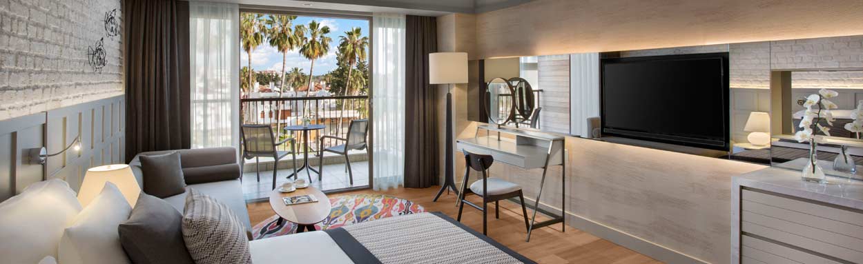Inside one of the hotel's suites, showing a large wall mounted television, a comfortable, large double bed, and cosy seating area. Beyond the full height patio doors is a furnished balcony, the room features neutral modern decor and all the facilities req