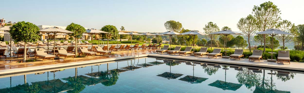 A view across the main swimming pool at the Anda Barut Collection hotel, with trees providing welcome shade behind the sun loungers positioned around the pool