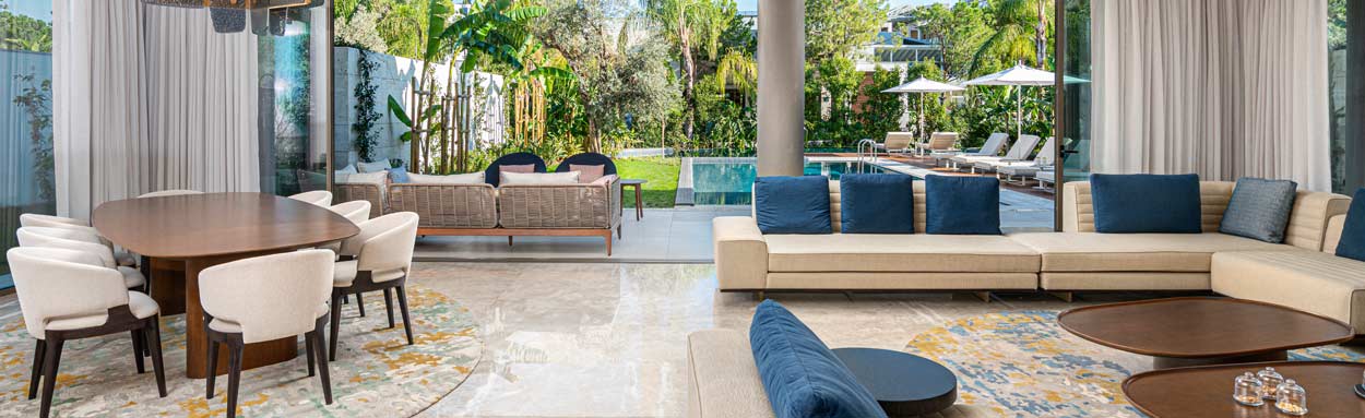 A large open plan villa with comfortable sofas and dining table extends out past floor to ceiling sliding doors to a landscaped garden that features a good size swimming pool with sun loungers, shade umbrellas and other garden furniture.