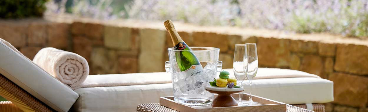 The Deluxe Collection offers the higest standard of service, accommodation and dining that Ikos offers. Images shows some sweet treats and a bottle of Tattinger Champagne in an ice bucket by the side of a sun lounger in the private garden of a Deluxe Coll