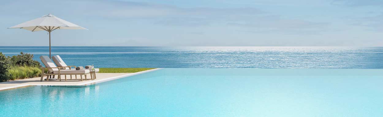 An inviting infinity swimming pool seems to blend seamlessly into the clear blue ocean, to the side of the pool sun loungers await guests at an Ikos Resort